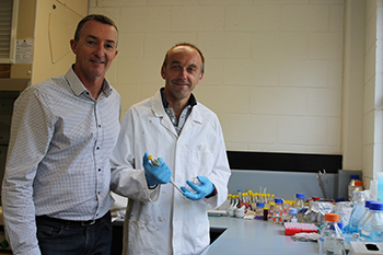 Two men stand next to a table in a laboratory. One is wearing a lab coat and gloves.